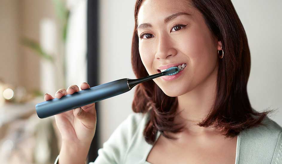 Een vrouw met halflang bruin haar gebruikt een donkerblauwe elektrische trimmer van het merk Philips bij haar hals, binnenshuis in zacht licht.