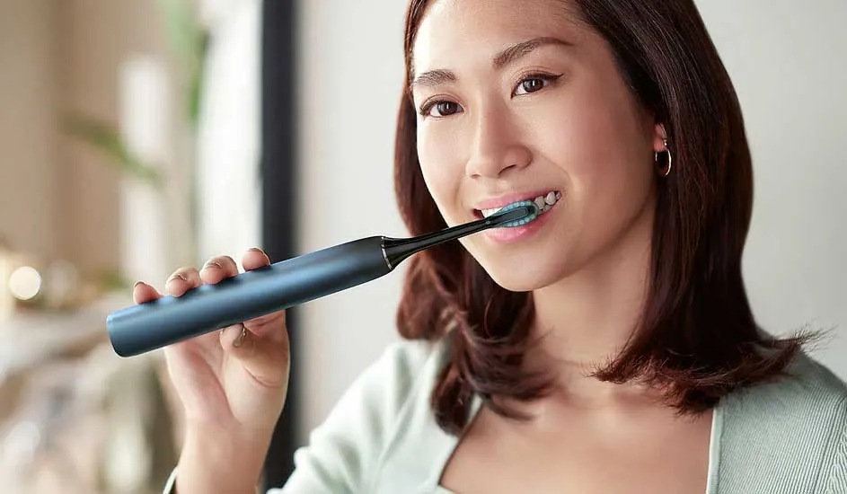 Een vrouw met halflang bruin haar gebruikt een donkerblauwe elektrische trimmer van het merk Philips bij haar hals, binnenshuis in zacht licht.