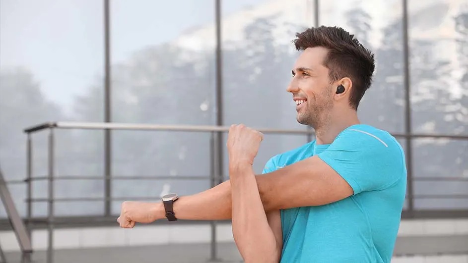 A sporty man in a blue top stretches his arm and smiles; he wears wireless earbuds and a fitness watch by a modern building.