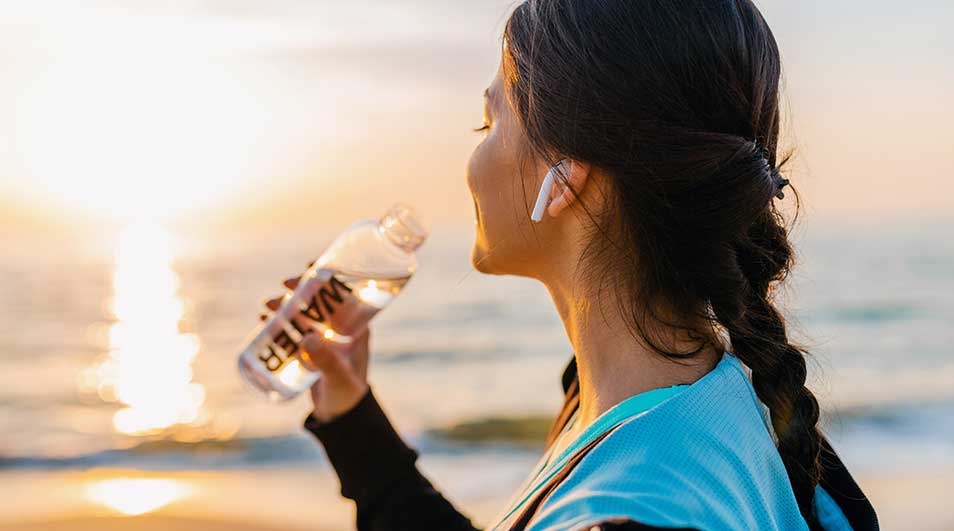 Vrouw met vlecht en draadloze oordopjes drinkt uit een fles water terwijl ze bij zonsondergang naar de zee kijkt. Op een warme avond buiten.