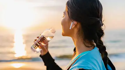 Vrouw met vlecht en draadloze oordopjes drinkt uit een fles water terwijl ze bij zonsondergang naar de zee kijkt. Op een warme avond buiten.