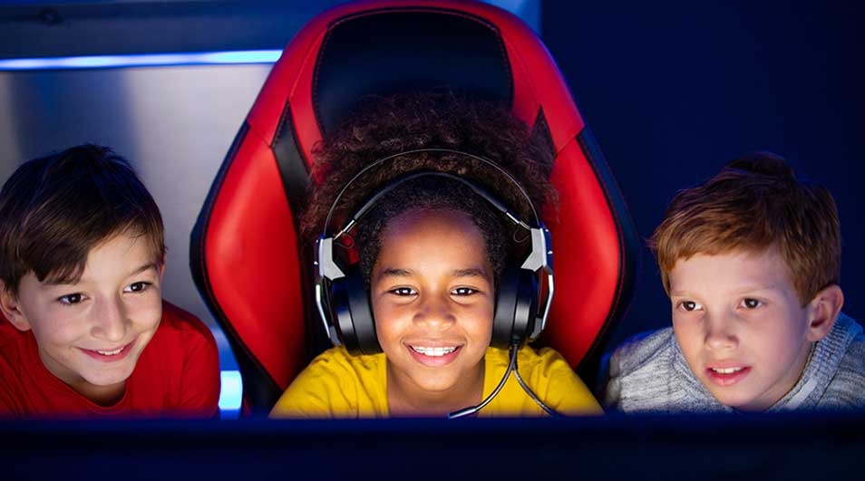 Three children sit together in front of a screen, one wearing a headset and sitting in a red game chair.