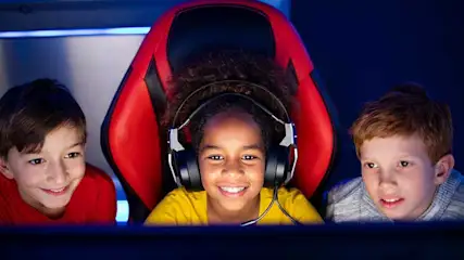 Three children sit together in front of a screen, one wearing a headset and sitting in a red game chair.