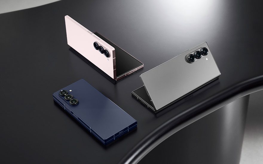 Drie Samsung Galaxy Z Fold smartphones in verschillende kleuren - roze, blauw en grijs - artistiek gerangschikt op donkere oppervlak met dramatische belichting.