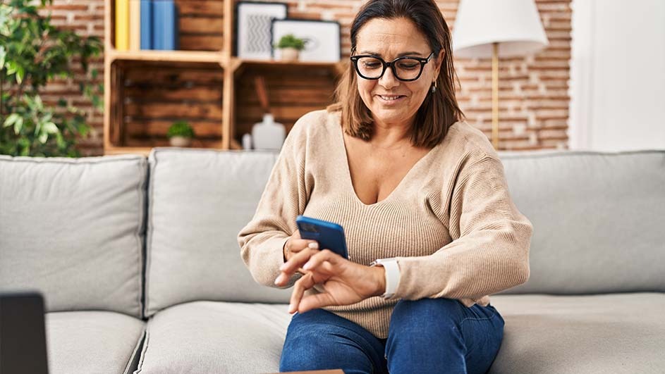 A woman wearing glasses and a tan sweater sits on a gray couch, holding a blue phone. A wooden shelf and brick wall are visible in the background.