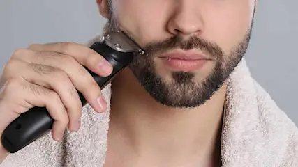 A person trims their beard with a black electric razor. A white towel is draped around their neck. The background is gray.