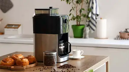 Koffiezetapparaat naast koffiebonen, kaneelbroodjes en een kopje op een houten tafel. Een groene plant staat op de achtergrond.