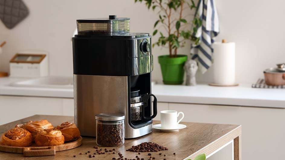 A modern coffee machine stands on a wooden kitchen island beside a jar of coffee beans, pastries, and a white cup; no brand name is visible.