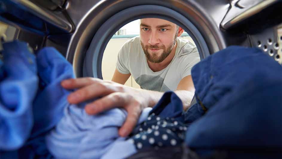 Man in a white shirt peers into a washing machine full of blue clothes, hand reaching in.