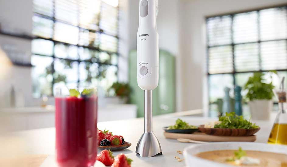 A white Philips hand blender stands on a bright kitchen counter, next to a berry smoothie, fresh herbs, salad and creamy soup.