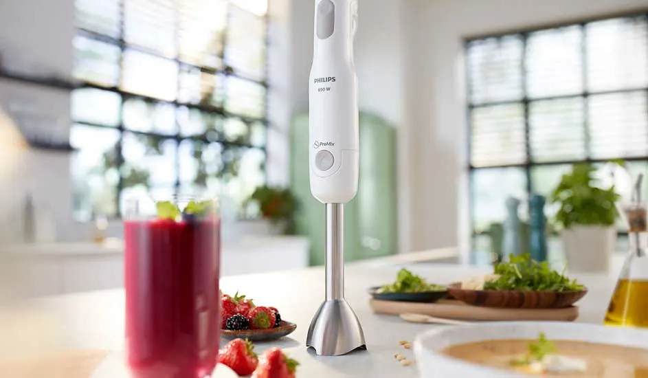 A white Philips hand blender stands on a bright kitchen counter, next to a berry smoothie, fresh herbs, salad and creamy soup.