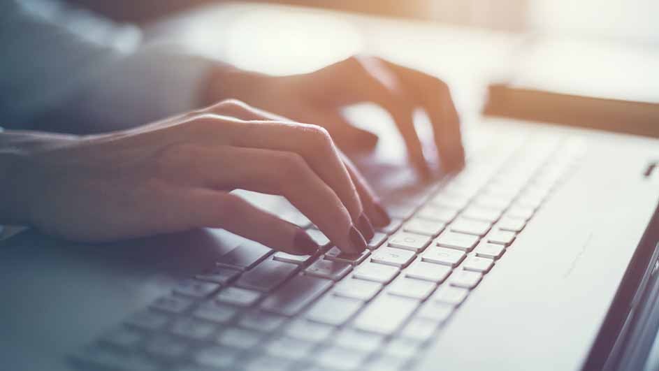 Hands are typing on a gray laptop keyboard. Fingers are pressing keys, with a bright background.