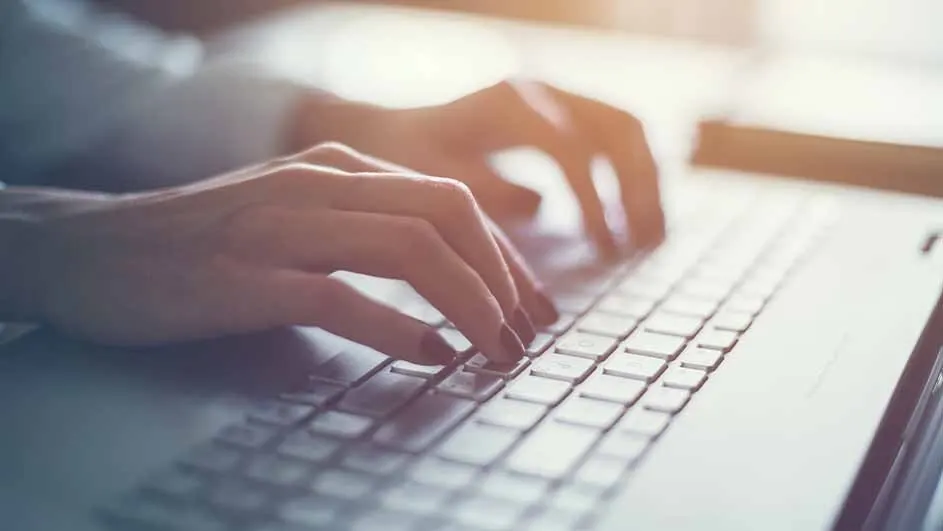 Hands are typing on a gray laptop keyboard. Fingers are pressing keys, with a bright background.