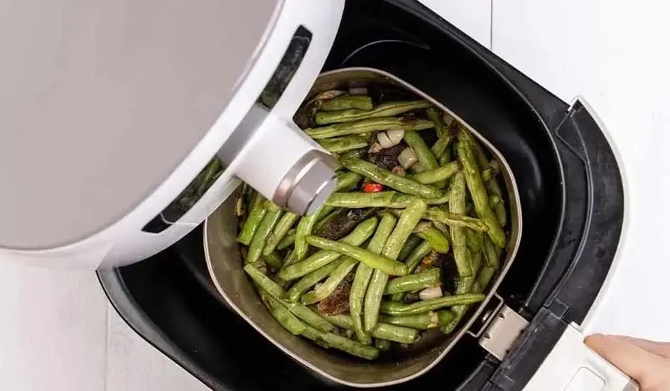Green beans with pieces of onion and pepper lie in the drawer of a white air fryer.