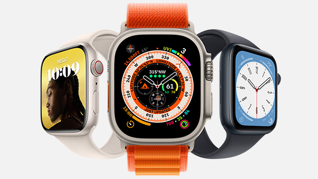Drie Apple Watches staan op een witte achtergrond. De middelste heeft een oranje band en een scherm met veel wijzers. De andere hebben een lichte en donkere band.