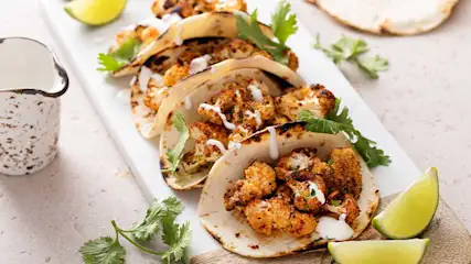 Taco's met bloemkool en limoen op een wit bord, met een kruik. Het gerecht is gegarneerd met koriander.