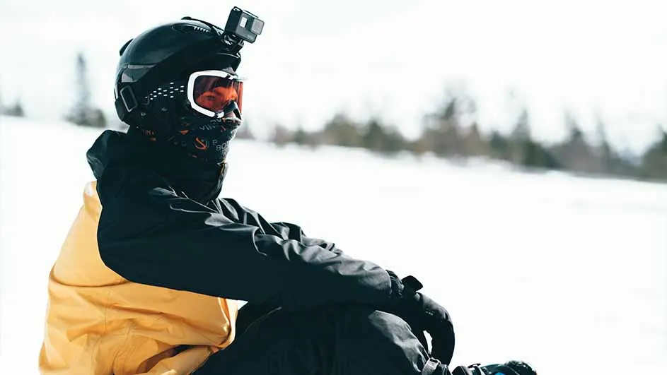 Een persoon in ski-uitrusting zit in de sneeuw met een zwarte helm, bril en een camera erop. De jas is zwart met geel.