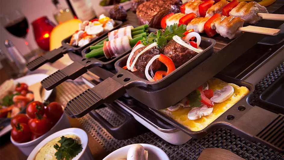 Een raclette grill met pannen met eten, waaronder vlees, groenten en kaas, omringd door kommen met verschillende ingrediënten.