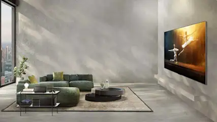 Een woonkamer met groene bank, ronde tafel op een vloerkleed en een tv met een afbeelding van dansers.