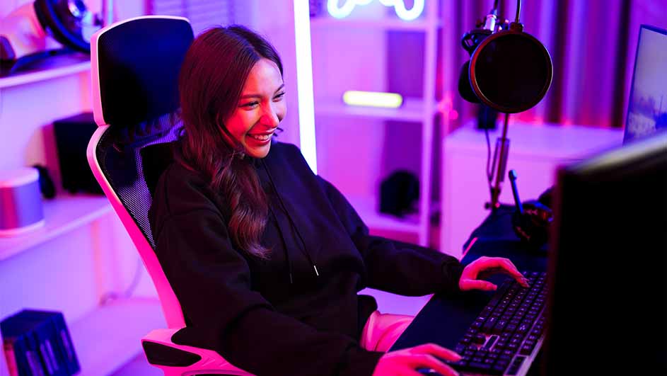Vrouw zit aan een bureau met een toetsenbord, microfoon en monitor, in een paars verlichte kamer met planken.