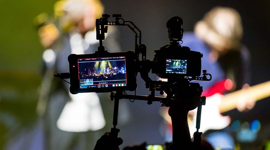Twee camera's filmen een live band op een podium. Schermen tonen de opname. Mensen en instrumenten vaag op de achtergrond.