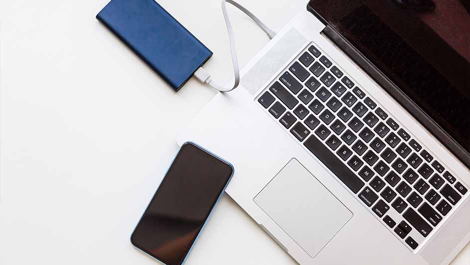Een witte tafel met een laptop, een blauwe powerbank met kabel en een zwarte smartphone.