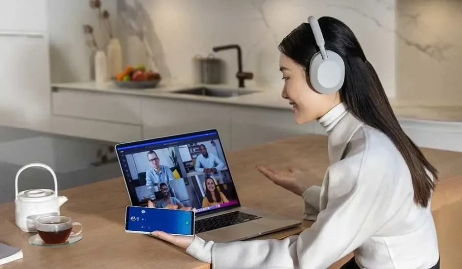 Een persoon met grijze koptelefoon voert een videogesprek op een Apple laptop en houdt een Samsung smartphone vast aan een keukentafel.