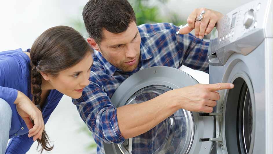A man in a blue plaid shirt points to the open door of a silver washing machine as a woman in blue looks on.