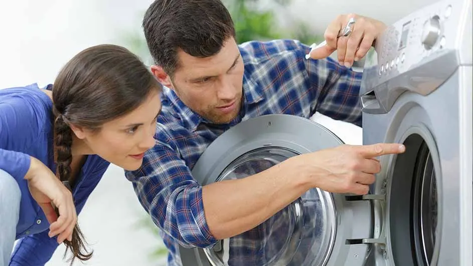 A man in a blue plaid shirt points to the open door of a silver washing machine as a woman in blue looks on.