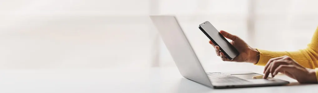A person with a yellow sweater uses a laptop and holds a smartphone in a bright room.
