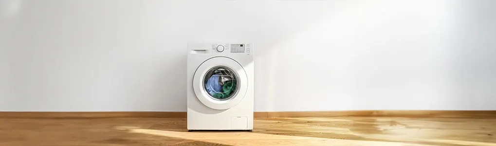 Een witte wasmachine staat op een houten vloer tegen een witte muur. Er zit gekleurde wasgoed in de trommel.