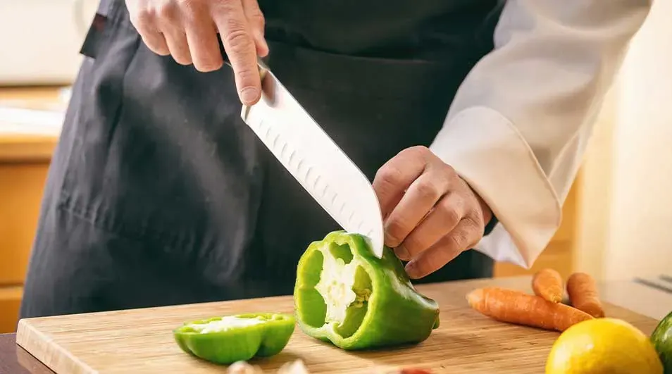 Een kok snijdt een groene paprika op een houten plank met een groot mes. Er liggen ook wortels en een citroen op de plank.