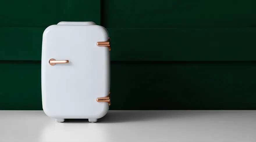 Een witte minikoelkast met koperkleurige handgrepen staat op een witte ondergrond tegen een donkergroene achtergrond.