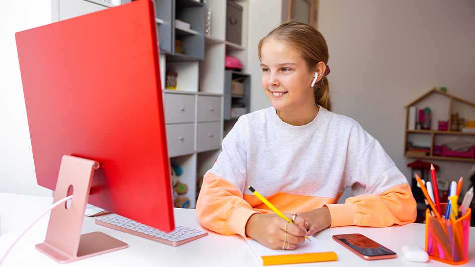 Meisje zit aan een bureau met een rode computer, een geel potlood en een smartphone. Op de achtergrond een witte boekenkast en een poppenhuis.