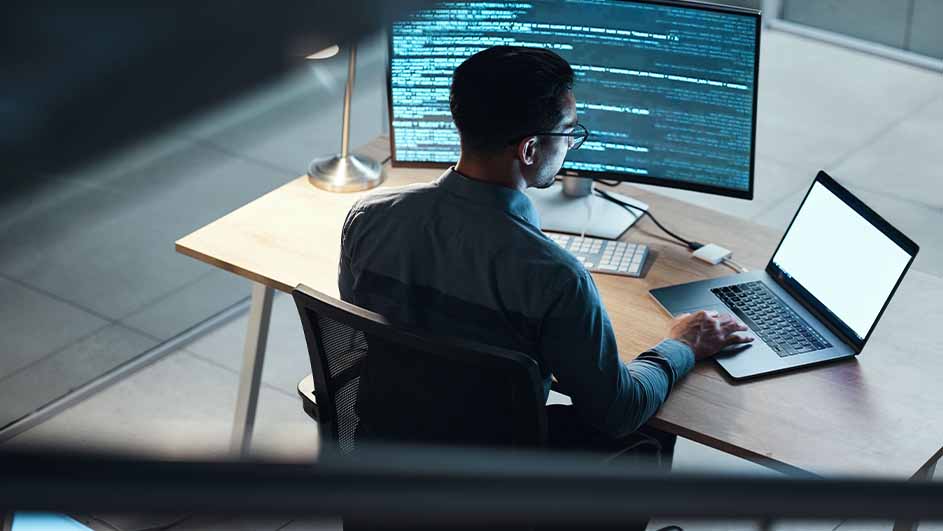 Een persoon zit aan een bureau met een computer, laptop en een lamp. Er staat code op het computerscherm.