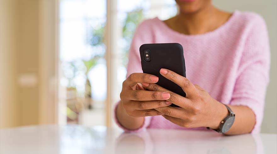 Een persoon met een roze trui houdt een zwarte smartphone vast met een horloge om de pols, tegen een vage achtergrond.