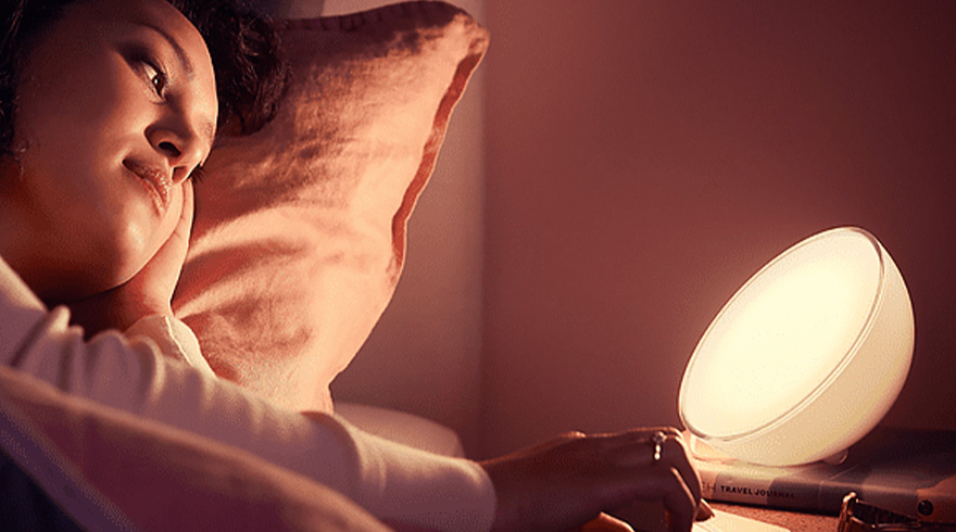 Vrouw leunt in bed met kussen, naast een witte lamp op een boek. De kamer is donkerrood verlicht door de lamp.