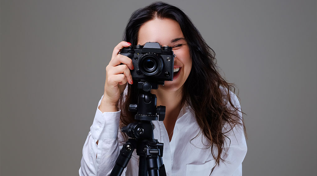 Een vrouw met een witte blouse gebruikt een zwarte camera op een statief tegen een grijze achtergrond.