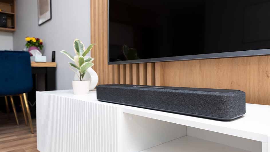 A soundbar sits on a white cabinet below a television, with a plant, flowers, and a blue chair in the background.