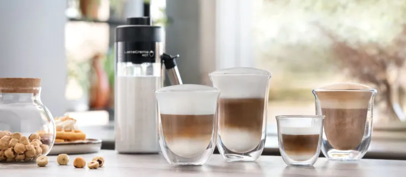 Verschillende glazen gevuld met koffie met melk en schuim, een melkopschuimer en een pot met hazelnoten op een tafel.
