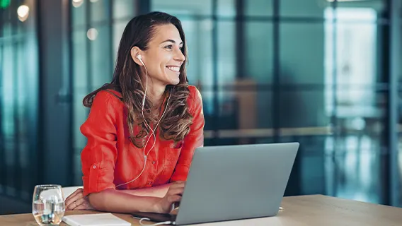 Vrouw met oortjes achter laptop aan tafel met glas water en notitieboekje, kijkend naar buiten. Op de achtergrond een kantoor.