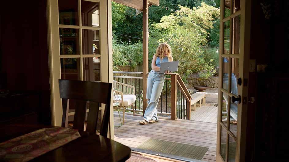Vrouw staat op houten terras met laptop, leunend tegen reling. Stoel en bank in de achtergrond. Openstaande deuren naar binnen.
