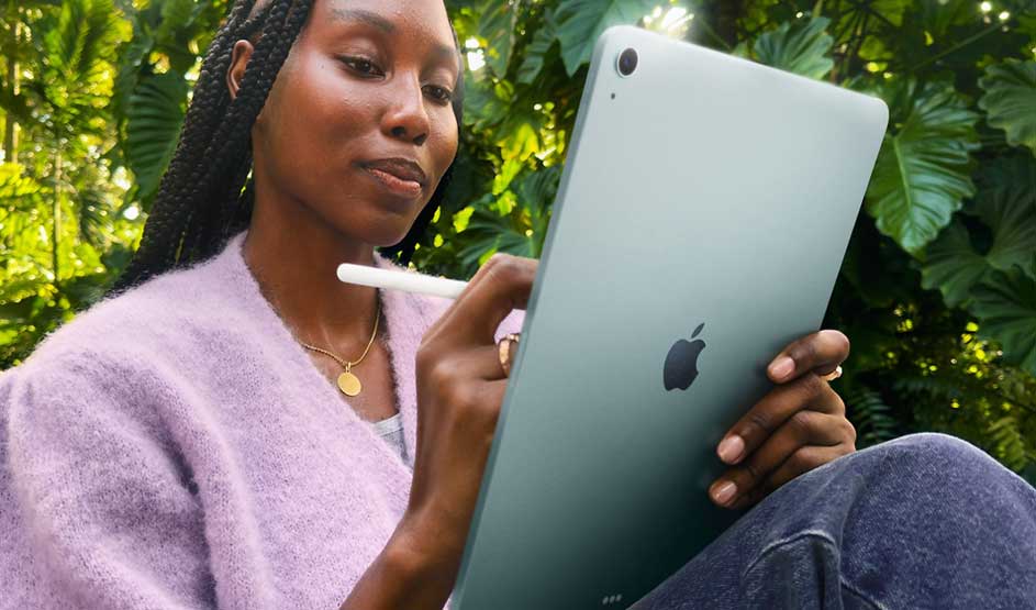 Iemand gebruikt een groene Apple iPad met een Apple Pencil, zittend buiten tussen groene planten en gekleed in een paarse trui.