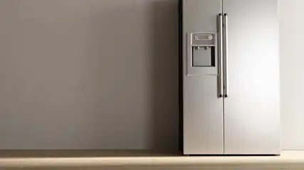 Een zilverkleurige koelkast met dubbele deuren en een waterdispenser staat op een houten vloer tegen een grijze muur.