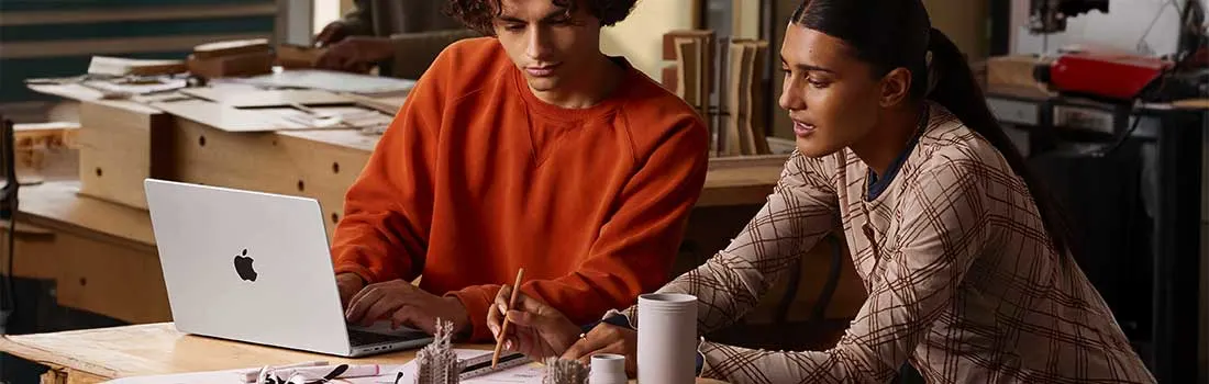 Twee studenten werken samen aan maquettes, terwijl één op de laptop typt en de ander schetst aan een drukke houten werktafel.