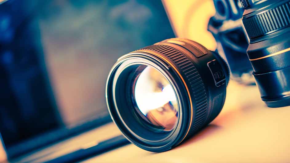Twee zwarte cameralenzen liggen op een bureau, met een laptop op de achtergrond.