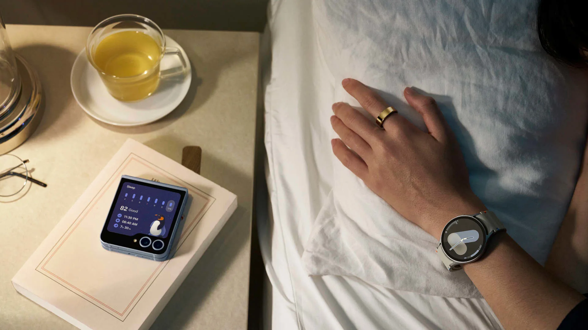 Nachtscène met telefoon op een boek, naast een glas sap en een hand met een horloge op een wit bed.