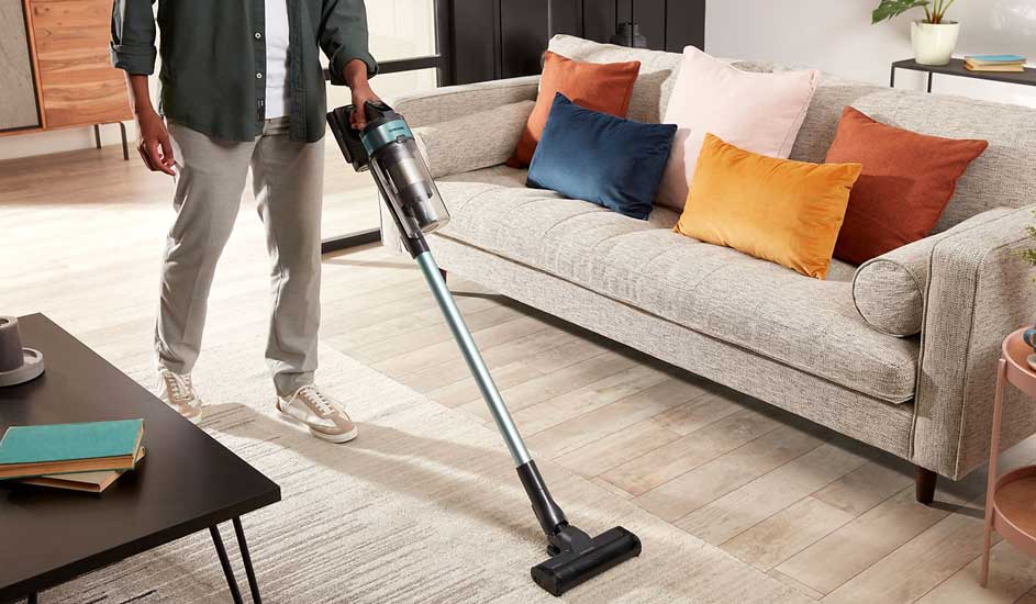 Een persoon stofzuigt een vloerkleed in de woonkamer met een draadloze Philips stofzuiger naast een bank met gekleurde kussens.