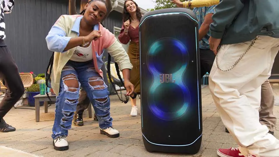 Mensen dansen rond een zwarte JBL-luidspreker met blauwe en groene lichten in een buitenomgeving.