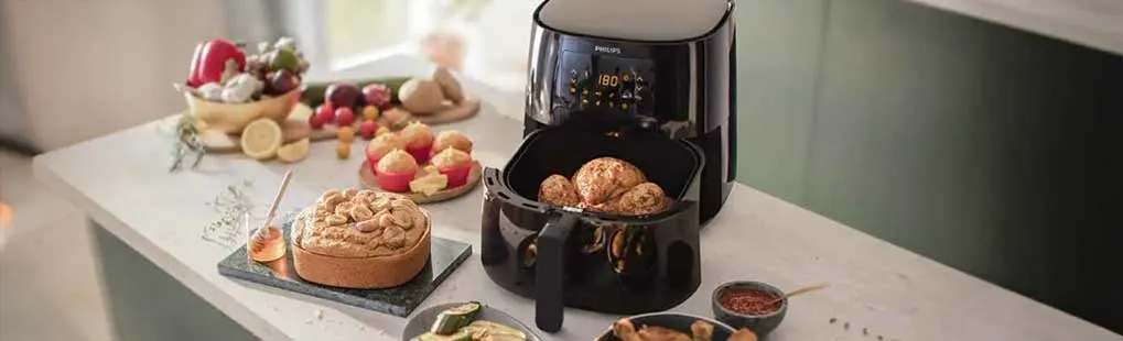 Een zwarte airfryer staat op een aanrecht omringd door gerechten zoals kippenpoten, cake en groenten.